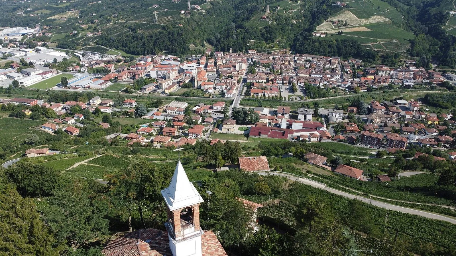 Santo Stefano Belbo visto dall'alto dalla collina del Moncucco, con il campanile di una chiesetta in primo piano, il paese nel fondovalle e le colline coperte di vigneti