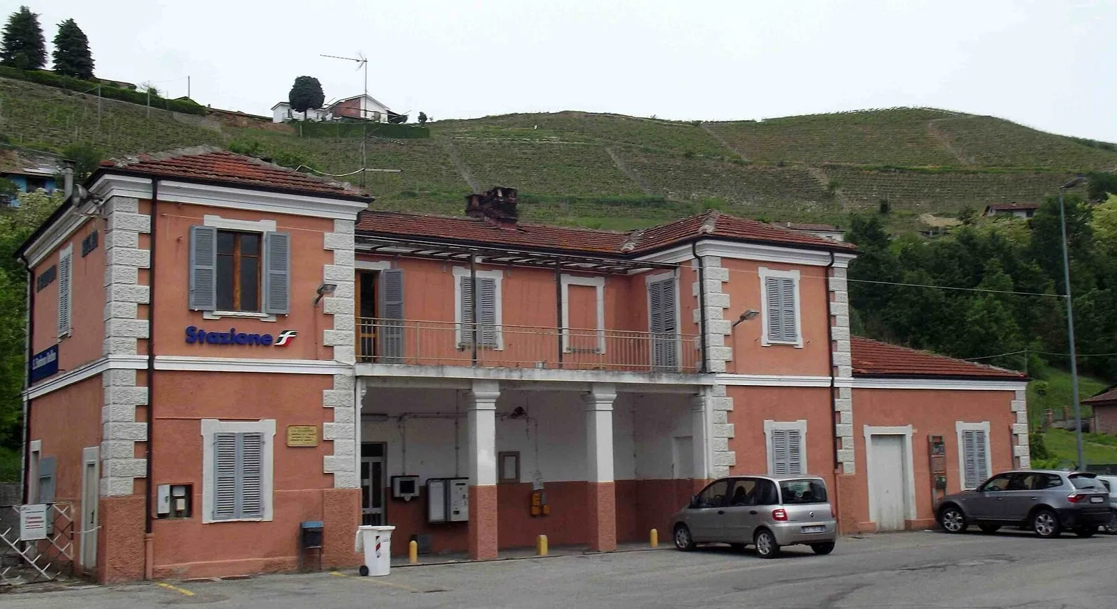 La stazione ferroviaria di Santo Stefano Belbo, edificio rosato con portico, e alle spalle una collina coltivata a vigneto